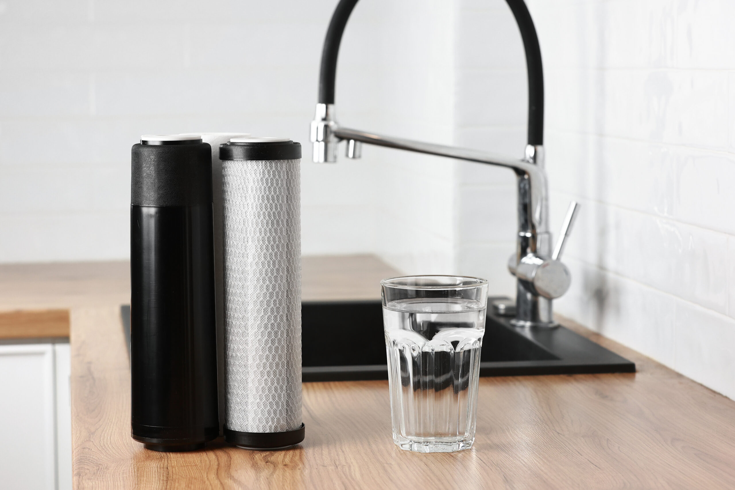 A glass of clean fresh water and set of filter cartridges on wooden table in a kitchen interior. Installation of reverse osmosis water purification system. Concept Household filtration system.